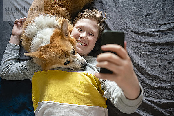 Porträt eines Jungen  der mit seinem Hund auf dem Bett liegt und mit seinem Smartphone Selfie nimmt