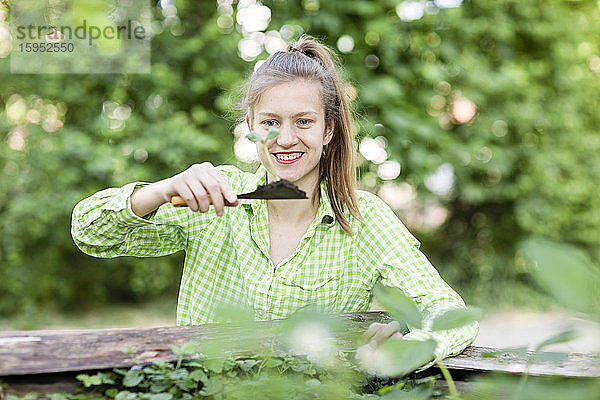 Lächelnde junge Frau betrachtet Sämling auf Kelle beim Pflanzen im Pflanzgefäß im Garten