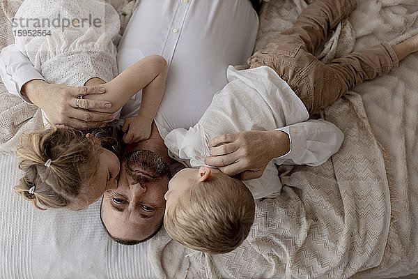 Porträt eines auf dem Bett liegenden Vaters  der von seinen beiden Kindern geküsst wird