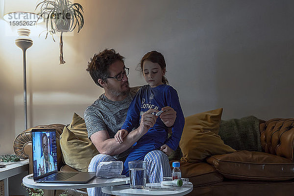 Mann hält Thermometer  während er mit seiner kranken Tochter während eines Videoanrufs zu Hause sitzt