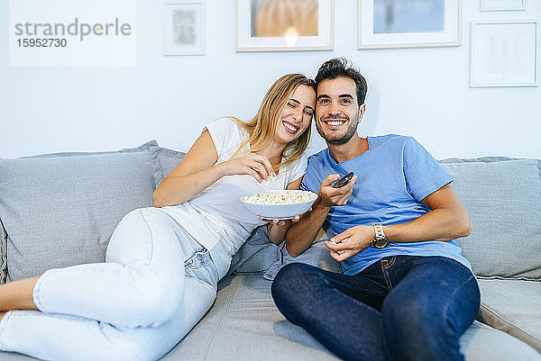 Lächelndes Paar beim Fernsehen  während es zu Hause auf dem Sofa Popcorn genießt