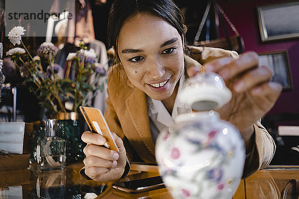 Neugierige Frau mit Kreditkarte betrachtet Vase im Secondhand-Laden