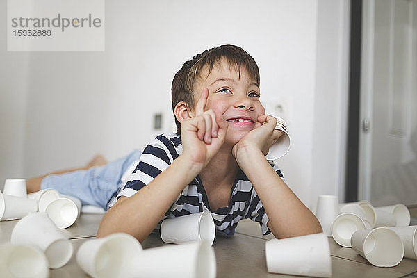 Porträt eines lächelnden kleinen Jungen  der zwischen weißen Pappbechern auf dem Boden liegt