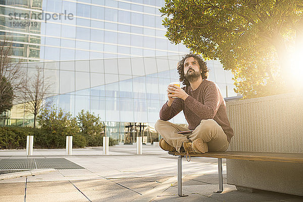 Porträt eines bärtigen Mannes  der sich bei Sonnenuntergang auf einer Bank mit Kaffee zum Mitnehmen ausruht