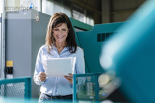 Lächelnde Geschäftsfrau bei der Durchsicht von Papieren in einer Fabrik
