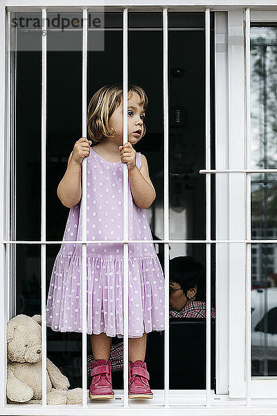 Mädchen steht am offenen Fenster und schaut durch das Fenstergitter
