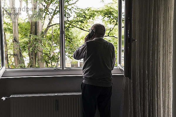 Rückenansicht eines älteren Mannes am Telefon  der am offenen Fenster steht