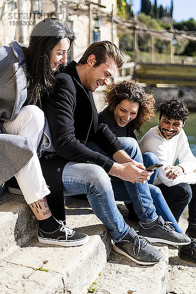 Vier glückliche Freunde sitzen auf einer Treppe am Gardasee und teilen sich ein Smartphone  Italien