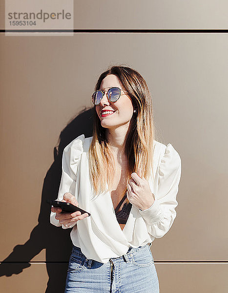 Porträt einer lächelnden jungen Frau mit Smartphone  die aus der Ferne betrachtet