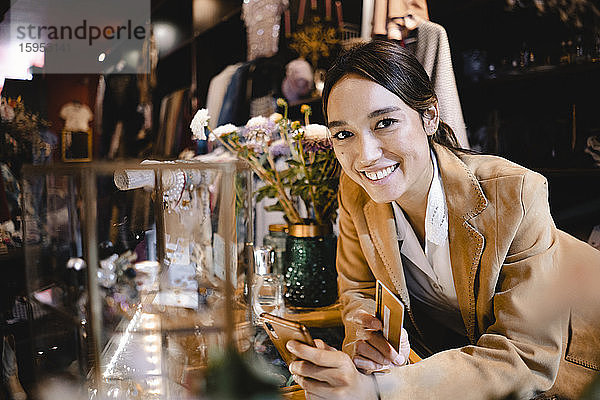 Lächelnde Frau mit Smartphone und Kreditkarte im Second-Hand-Laden