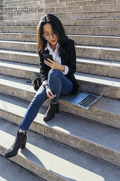 Junge Geschäftsfrau mit Laptop sitzt auf einer Treppe im Freien und schaut auf ihr Handy