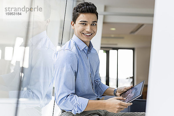 Porträt eines lächelnden jungen Geschäftsmannes mit Tablette am Fenster im Büro
