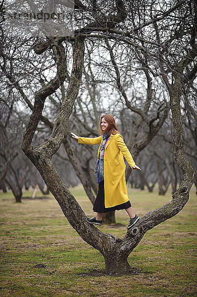 Frau in gelbem Mantel in voller Länge auf nacktem Baum im Park stehend