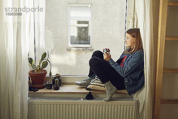 Mädchen sitzt zu Hause mit Smartphone am Fenster