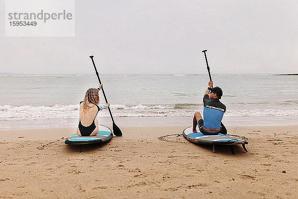 Rückansicht des Ausbilder und der jungen Frau auf Paddelbrettern mit Rudern am Strand sitzend