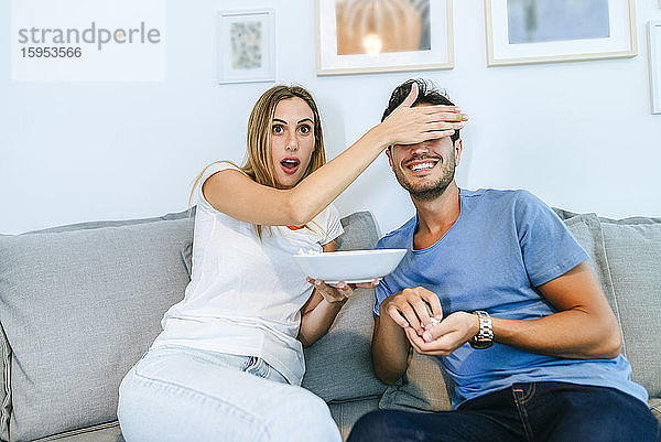 Geschockte Frau bedeckt die Augen des Mannes  während sie zu Hause im Wohnzimmer fernsieht