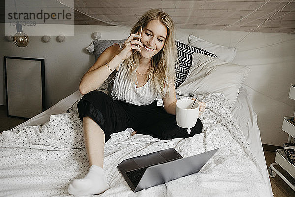 Junge Frau mit Laptop trinkt etwas  während sie im Schlafzimmer zu Hause mit dem Handy telefoniert