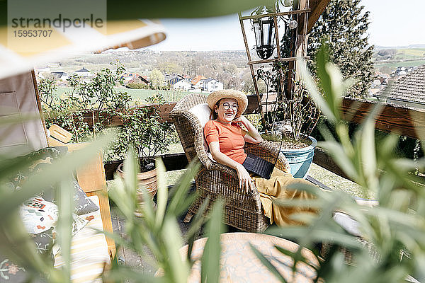 Glückliche reife Frau sitzt auf der Terrasse mit Laptop auf dem Schoß und entspannt