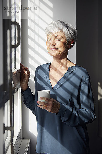 Porträt einer älteren Geschäftsfrau mit Kaffeetasse  die am Fenster steht und das Sonnenlicht genießt