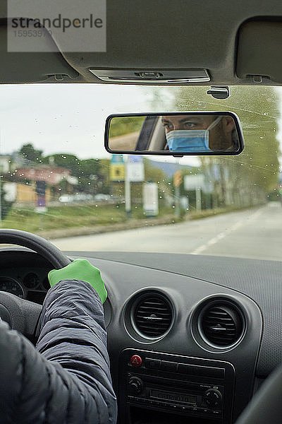 Mann mit Maske und Handschuhen fährt Auto und reflektiert im Rückspiegel in der Stadt