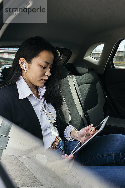 Junge Geschäftsfrau sitzt im Auto und benutzt Ohrstöpsel und digitales Tablet