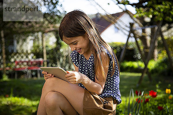 Glückliches Mädchen sitzt im Garten und benutzt digitales Tablett