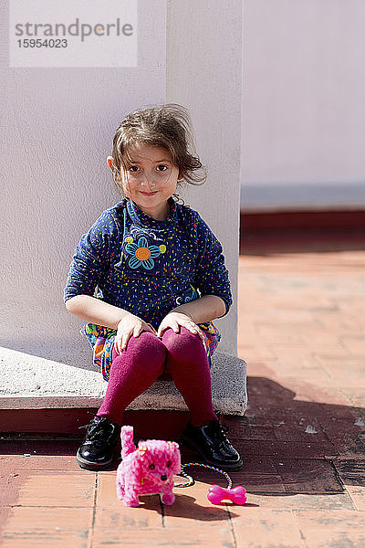 Porträt eines lächelnden kleinen Mädchens  das mit seinem rosa Spielzeughund auf dem Dach sitzt