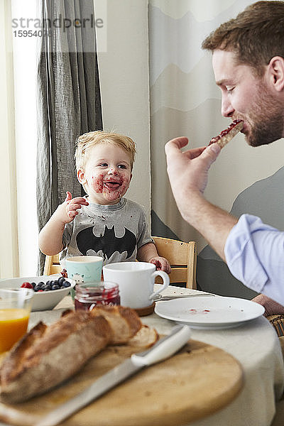 Porträt eines verschmierten kleinen Jungen am Frühstückstisch  der seinen Vater beobachtet  wie er Brot mit Marmelade isst