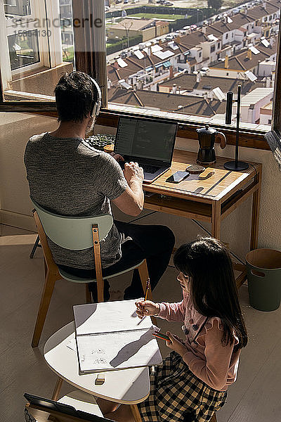Vater arbeitet am Laptop  während seine Tochter Hausaufgaben macht