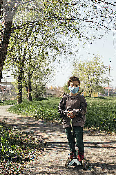 Porträt eines Jungen mit Schutzmaske mit Kickroller in einem Park