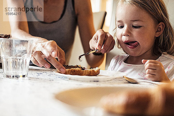 Porträt eines kleinen Mädchens  das seiner Mutter dabei zusieht  wie sie Schokoladencreme auf ein Croissant streicht