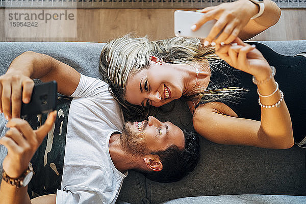 Glückliches Paar liegt auf der Couch und nimmt sich mit Smartphones ein  während es sich gegenseitig ansieht
