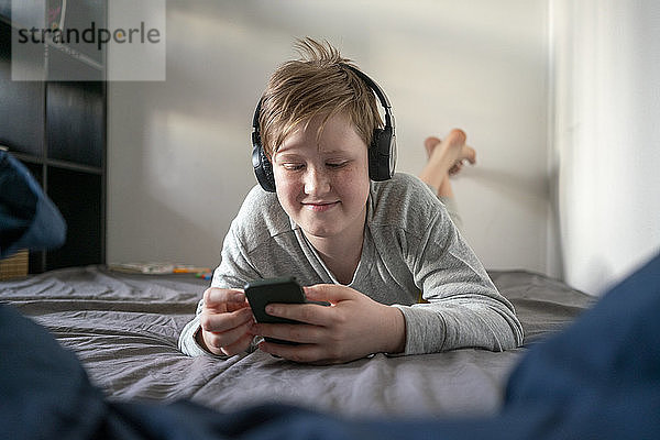 Porträt eines lächelnden Jungen mit Kopfhörern  der auf dem Bett liegt und auf sein Handy schaut