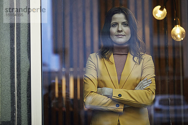 Porträt einer selbstbewussten Geschäftsfrau im gelben Anzug hinter einer Fensterscheibe stehend
