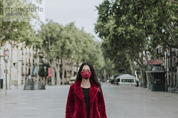 Porträt einer Frau mit roter Gesichtsmaske und Jacke  die auf einer leeren Straße in der Stadt steht  Barcelona  Spanien