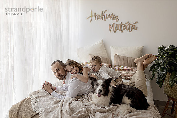 Porträt eines auf dem Bett liegenden Vaters mit seinen beiden Kindern und seinem Hund