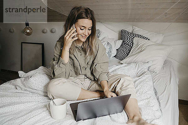 Lächelnde Frau spricht mit einem Smartphone  während sie zu Hause im Schlafzimmer am Laptop arbeitet