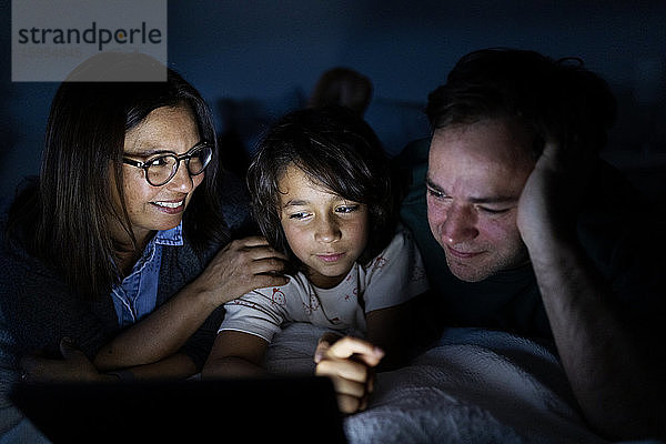 Eltern und Sohn liegen zu Hause gemeinsam auf dem Bett und verwenden ein digitales Tablett