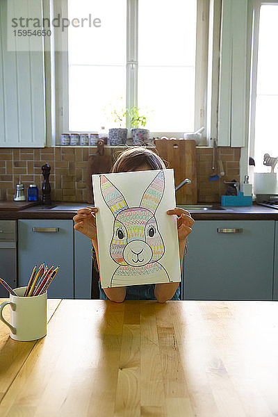 Kleines Mädchen sitzt am Küchentisch und hält eine Zeichnung des Osterhasen vor dem Gesicht