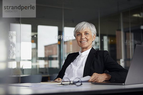 Porträt einer lächelnden älteren Geschäftsfrau  die im Büro am Schreibtisch sitzt