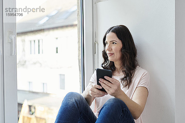 Nachdenkliche Geschäftsfrau mit Smartphone schaut durch das Fenster im Büro
