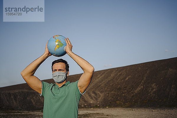 Erwachsener Mann mit Maske  der einen Globus auf einer stillgelegten Minenspitze hält