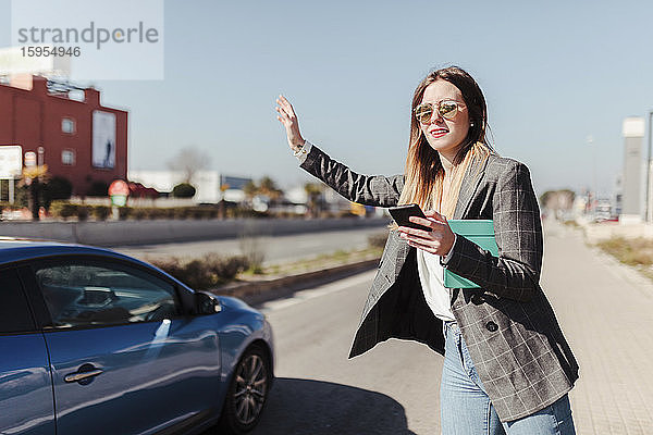Junge Geschäftsfrau mit digitalem Tablet und Smartphone steht am Straßenrand und ruft Taxi