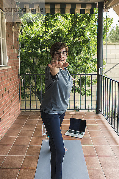 Ältere Frau praktiziert Yoga mit Laptop auf dem Balkon