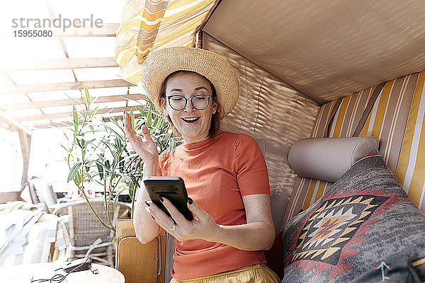 Porträt einer aufgeregten reifen Frau  die auf der Terrasse sitzt und auf ein Smartphone schaut