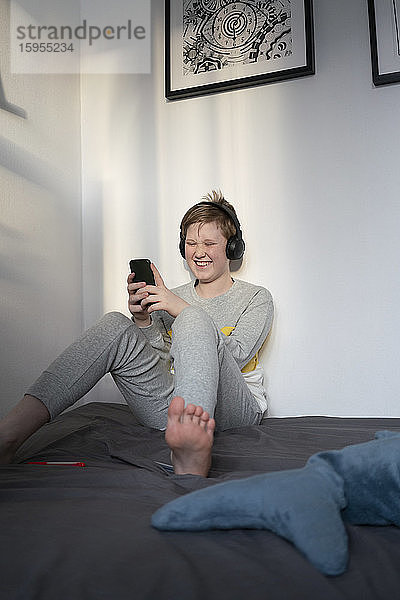 Porträt eines lachenden Jungen  der mit Kopfhörern auf dem Bett sitzt und auf sein Handy schaut