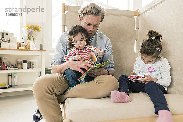Vater liest der Tochter ein Buch vor  während ihre Schwester spielt