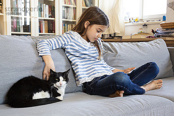 Mädchen sitzt auf Couch mit digitalem Tablett und streichelt Katze