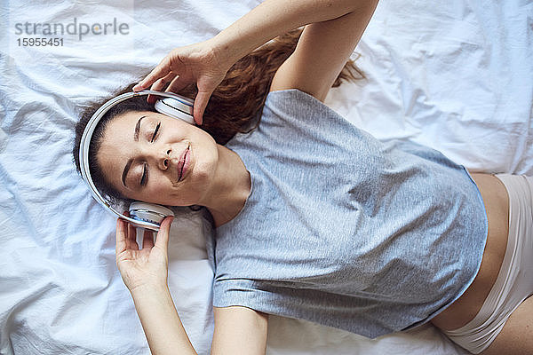 Porträt einer jungen Frau mit geschlossenen Augen auf dem Bett liegend und mit Kopfhörern Musik hörend