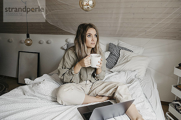 Nachdenkliche Frau mit Laptop  die zu Hause am Bett sitzt und Kaffee trinkt